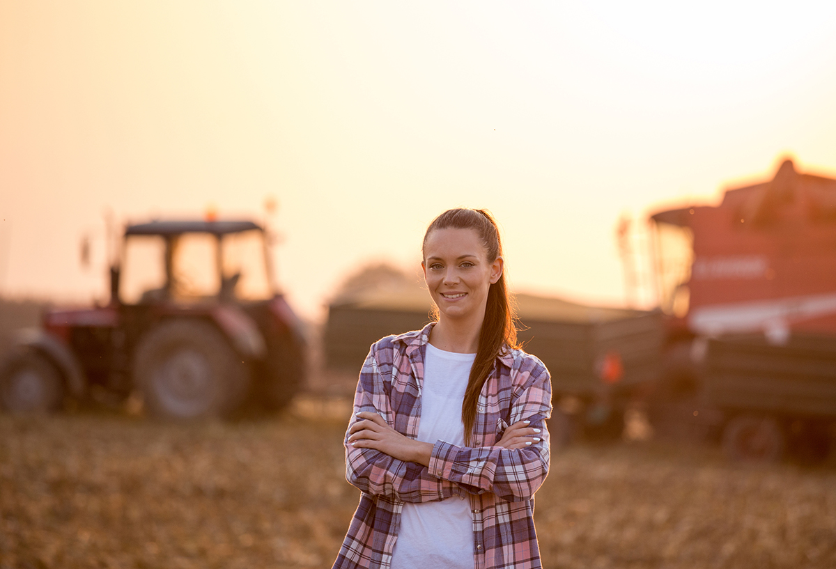 Satisfied young farmer woman standing with crossed arms in front of combine harvester and tractor with trailer in corn field